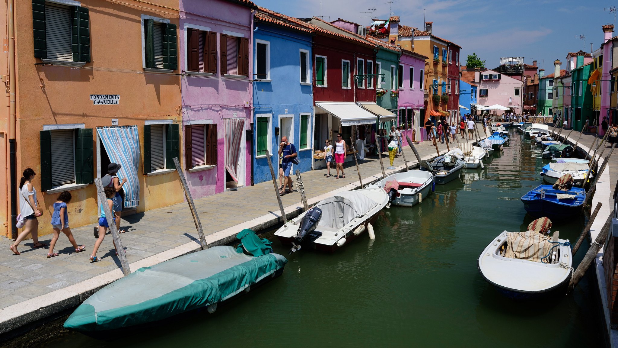 A canal on Burano lined with houses painted in flat blue, orange, red and green, with boats moored along it