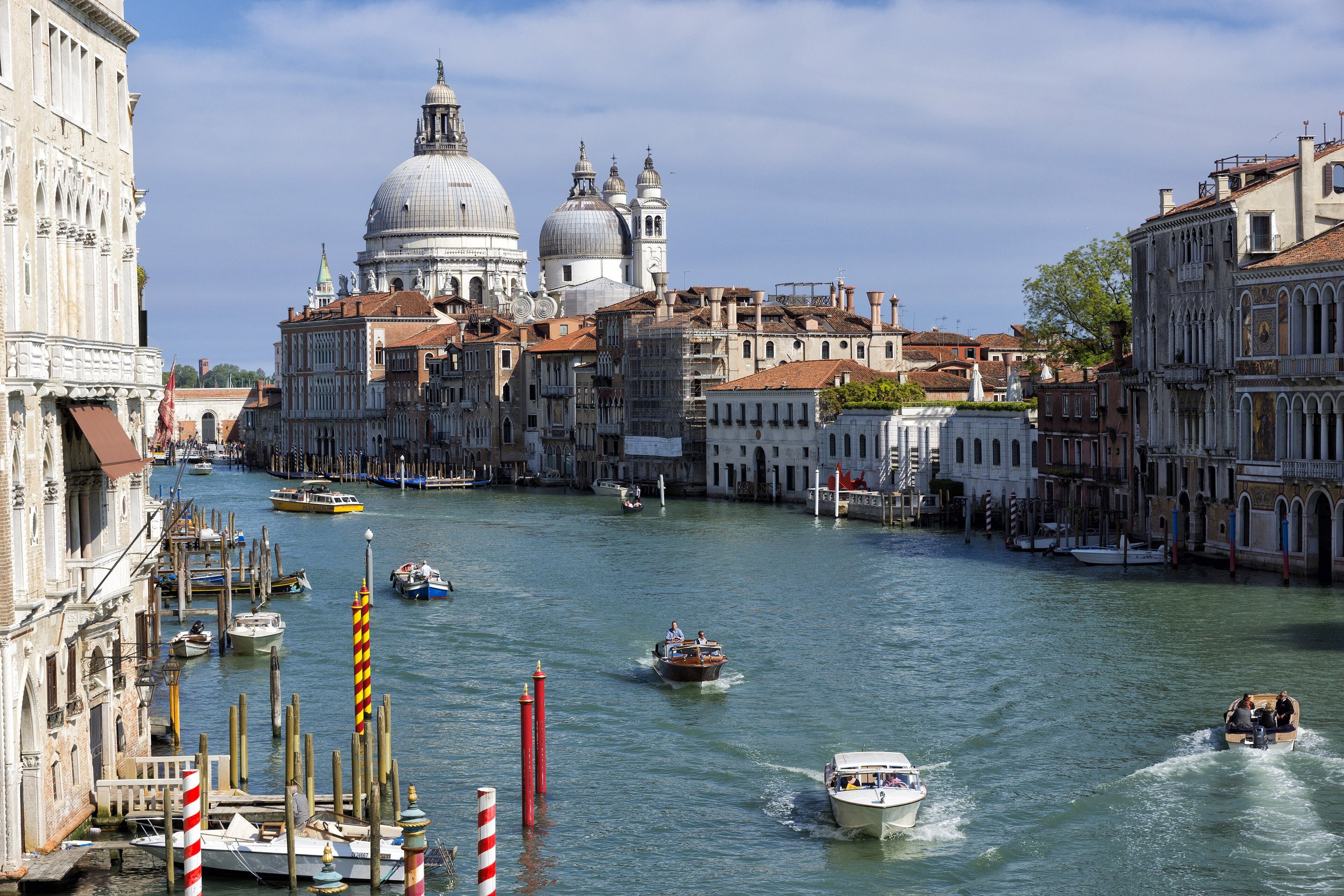 The Grand Canal in Venice with a cluster of striped bricole — the oak mooring posts that mark the navigable channel — standing in the water in the foreground, palazzi and boat traffic behind them