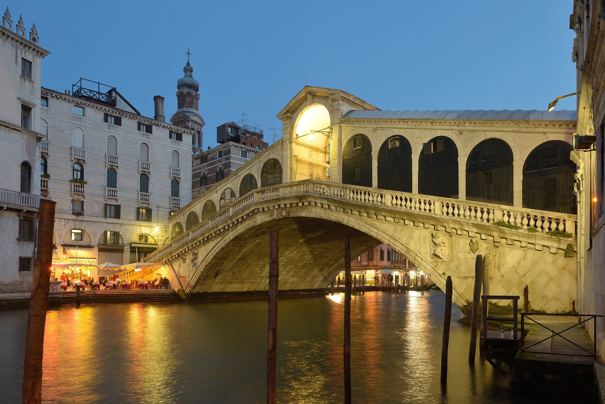 The Rialto Bridge lit at dusk from the west, its arch and shop arcade reflected in the Grand Canal