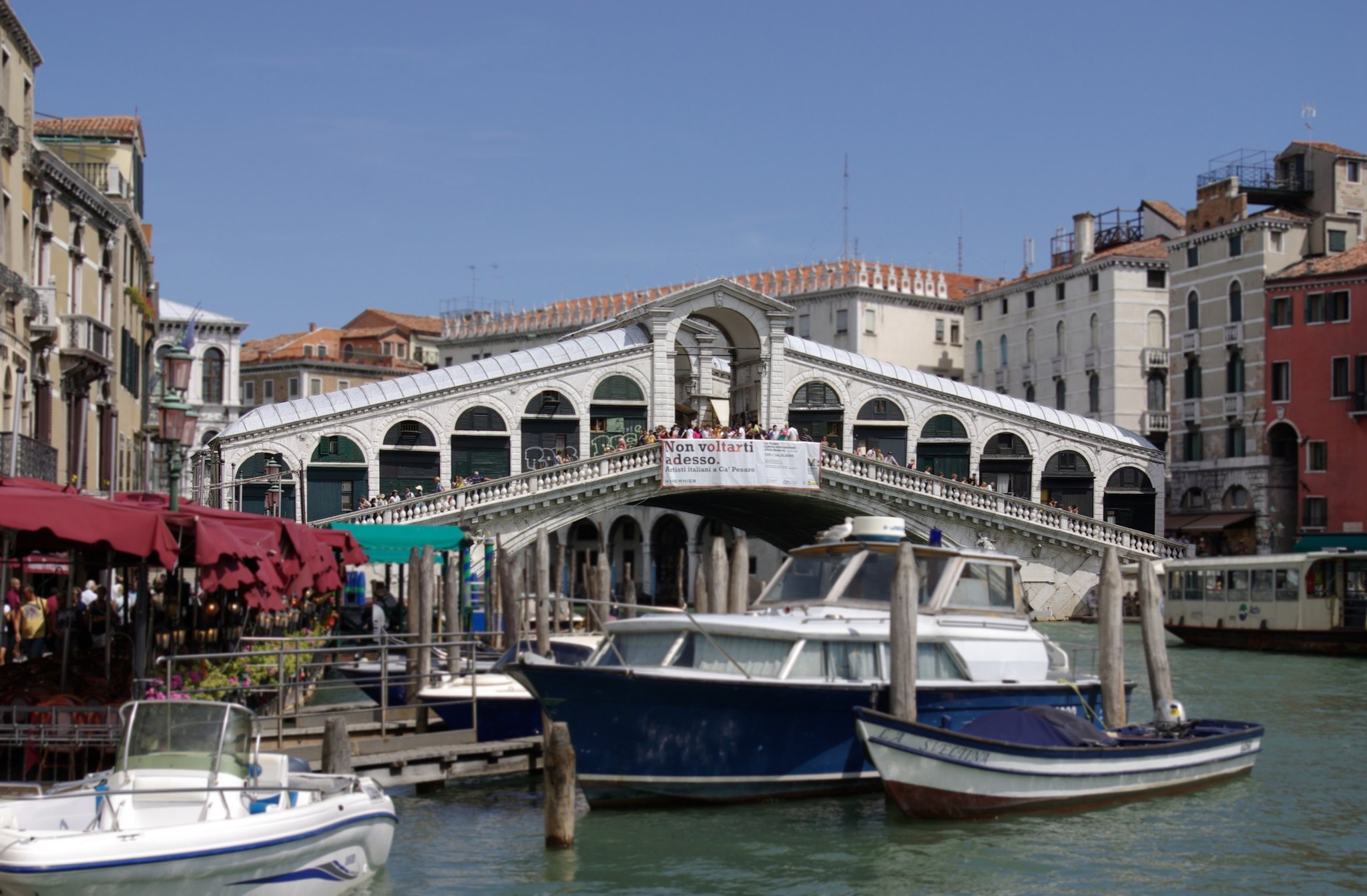 The Rialto Bridge in daylight, boats and canalside awnings crowding the water beneath it